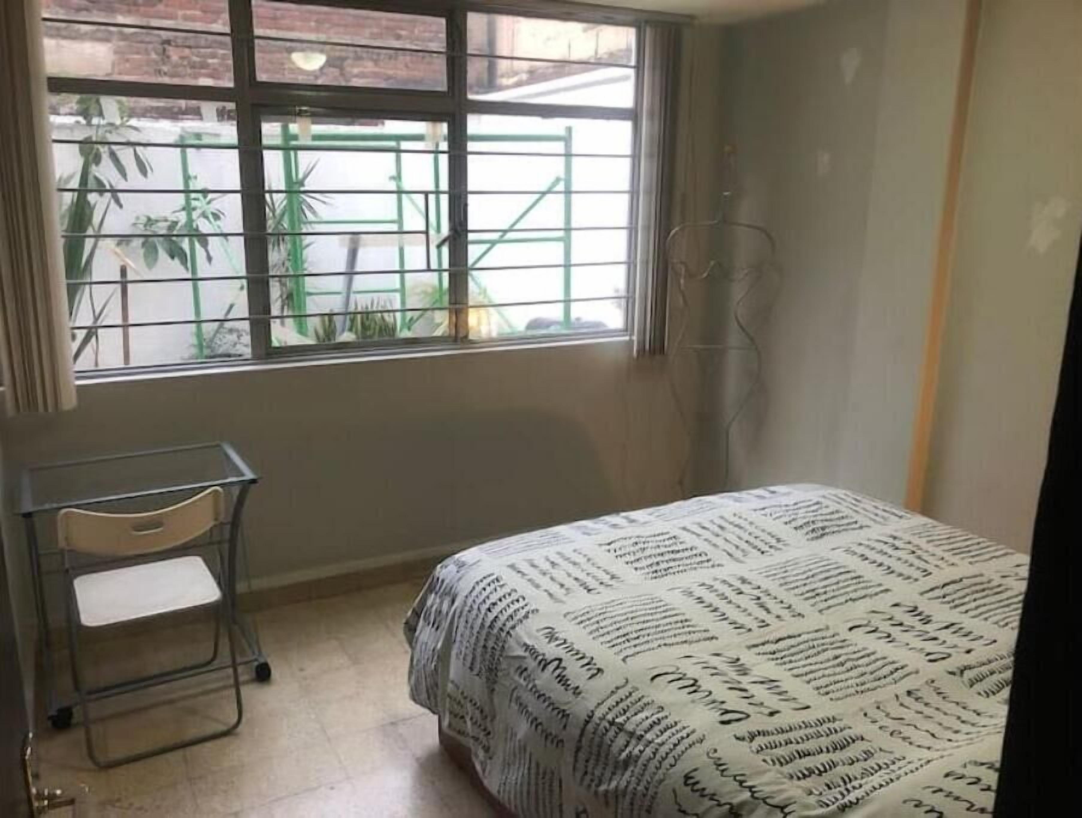 A bedroom with a bed, a desk, and a chair. There is a window with a view of greenery outside.