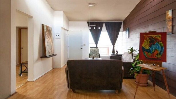 A modern living room with a dark sofa, a small table, and a potted plant. There is a painting on the wall and a window with curtains. - Sunset Included in your stay !!! (Tijuana)