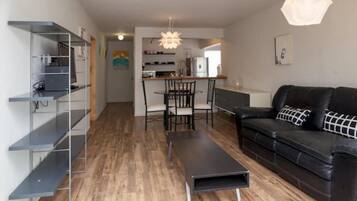 Un salon moderne avec un canapé en cuir noir, une table à manger avec des chaises, et un coin cuisine à l’arrière-plan.