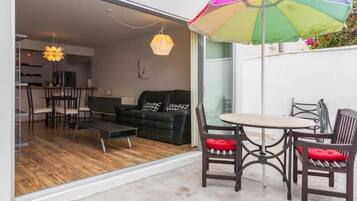 Un salon moderne avec un canapé, une table basse et des fauteuils, une porte ouverte donnant sur un coin repas, et un parapluie coloré à l’extérieur.