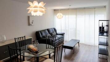 Un salon moderne avec une table à manger en verre, un canapé en cuir noir et une grande fenêtre équipée de stores.