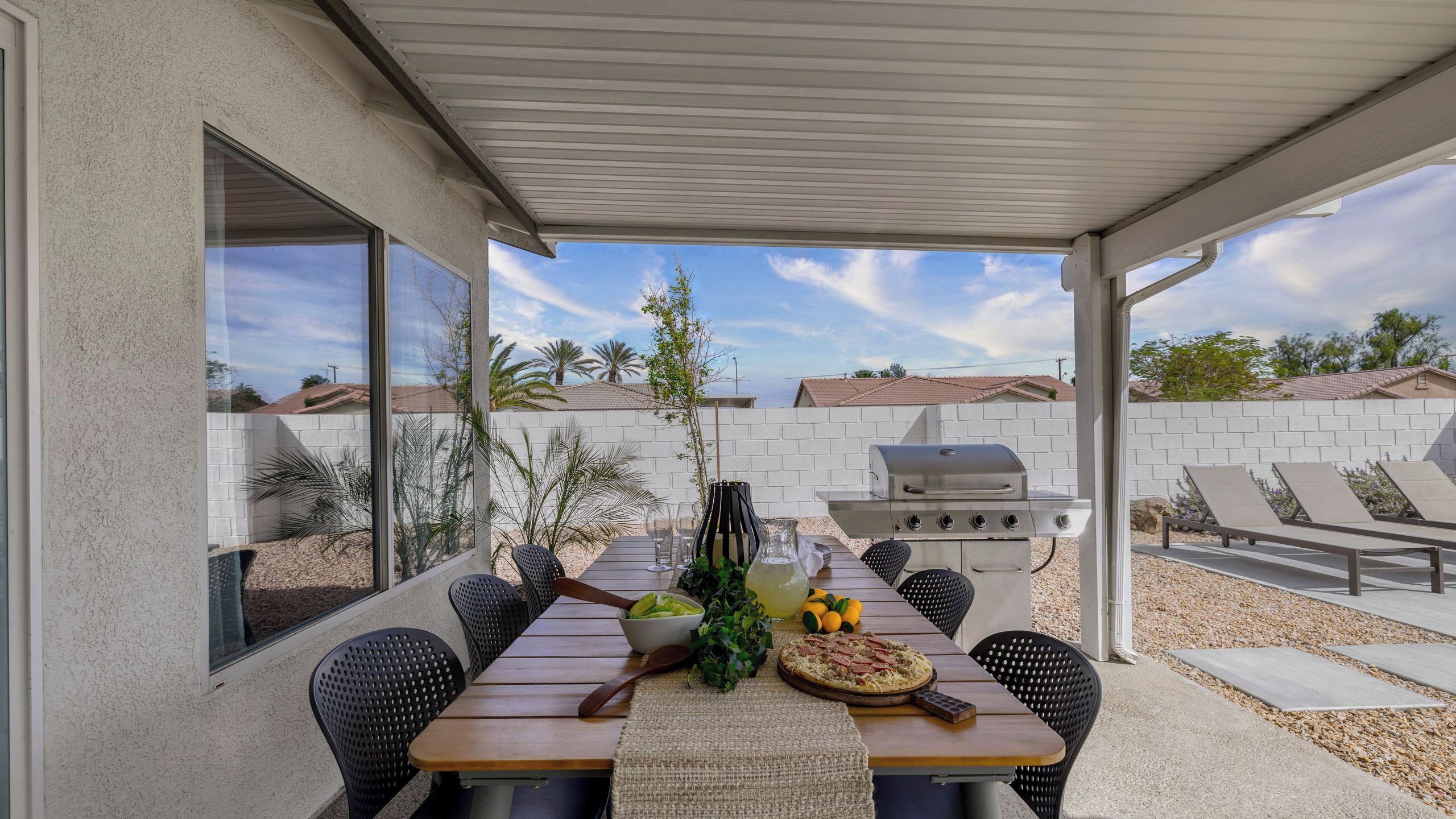 An outdoor patio with a wooden table, chairs, a grill, and a view of the neighborhood.