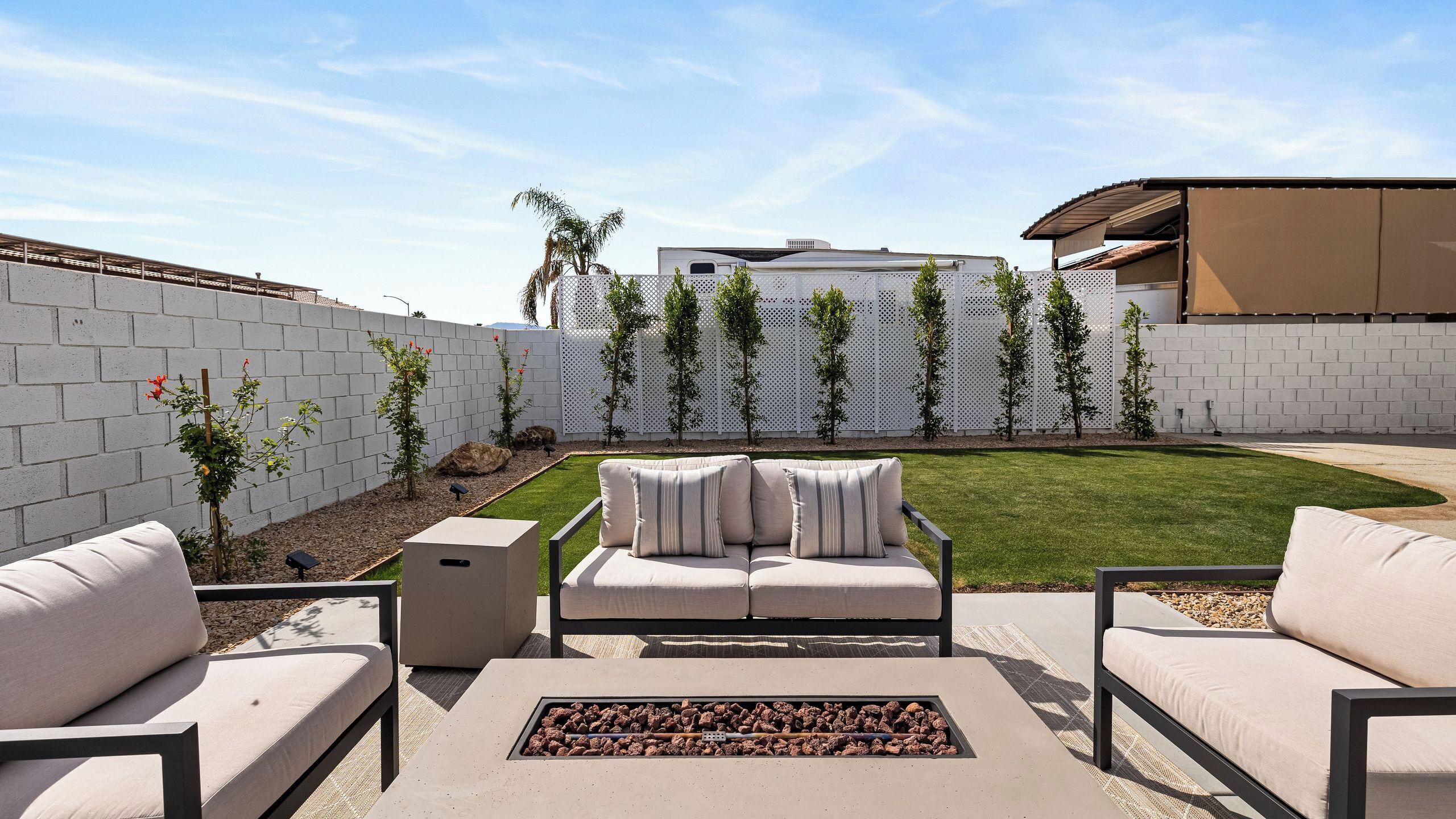 A patio with outdoor furniture, a fire pit, and a white fence with trees.