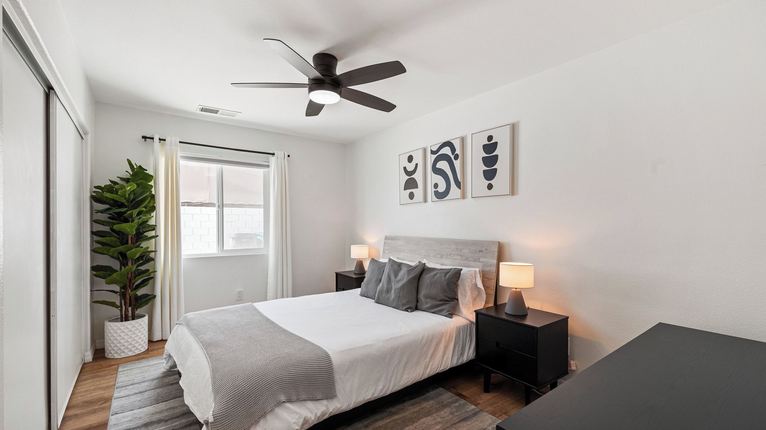 A bedroom with a bed, bedside tables, a ceiling fan, a window, and artwork on the wall.