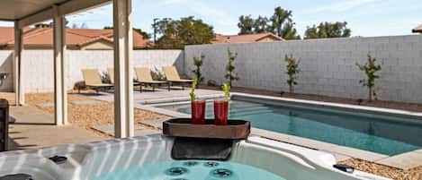 A hot tub with a tray holding two drinks, a pool, and a patio area with lounge chairs and a white brick wall.