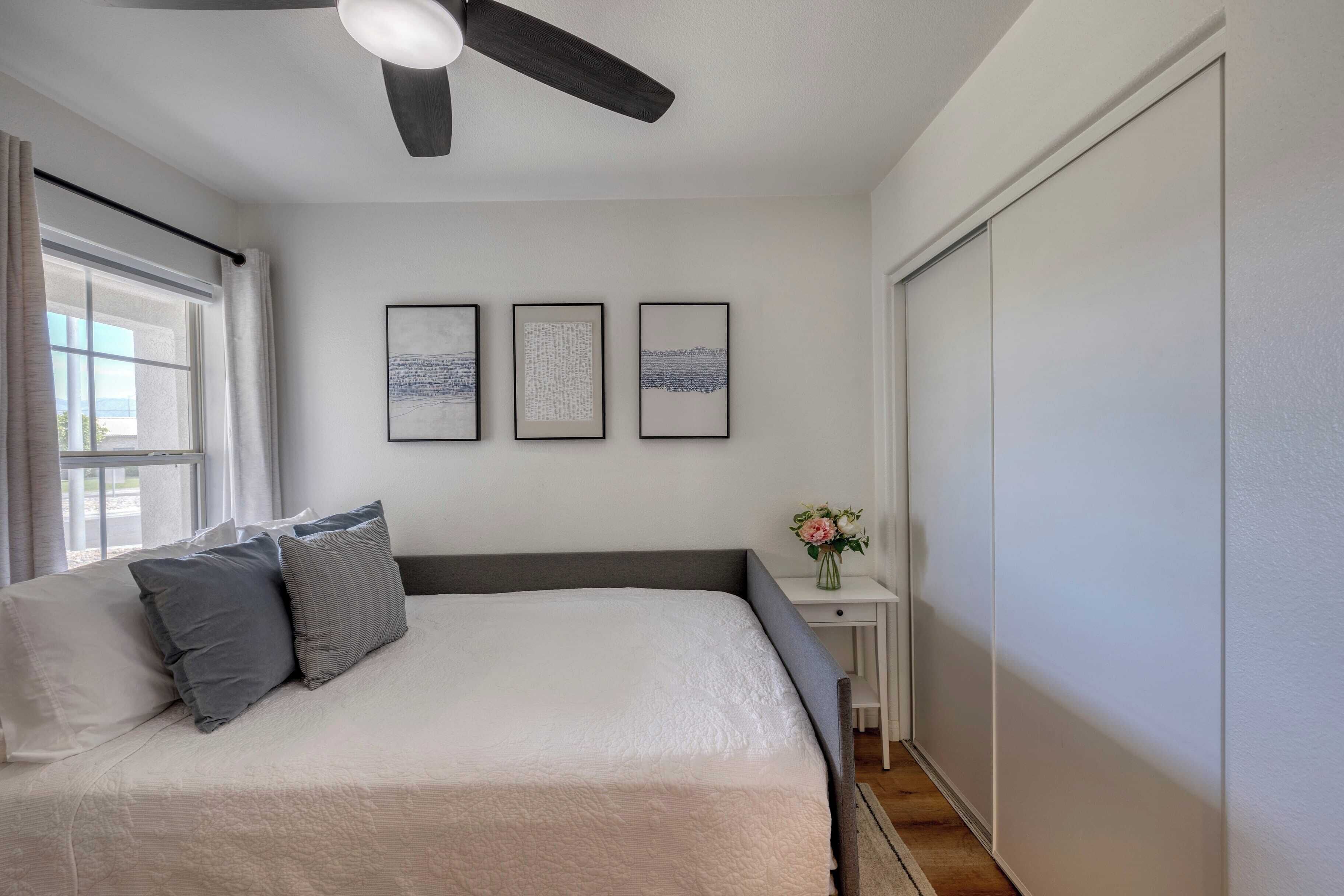A bedroom with a bed, a ceiling fan, a window with curtains, and three framed pictures on the wall.