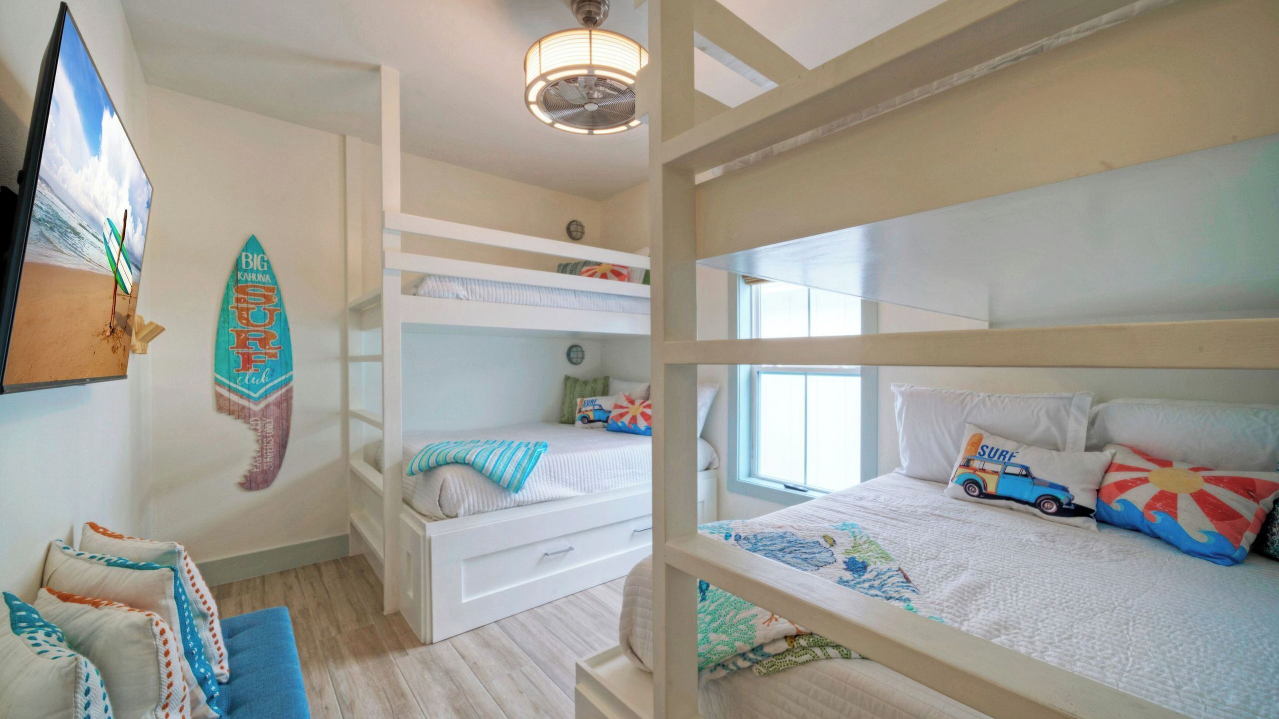 Une chambre avec des lits superposés, un ventilateur de plafond, une décoration sur le thème du surf et une fenêtre laissant entrer la lumière naturelle.