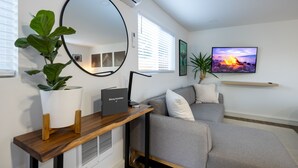 Uma sala de estar moderna com um sofá cinzento, uma mesa lateral de madeira, um espelho grande e uma TV exibindo uma paisagem de pôr do sol.