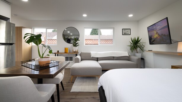 Quarto moderno e bem iluminado com área de refeições, sofá, cama e televisão na parede.
