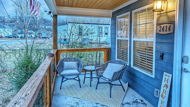 A covered porch with two wicker chairs and a small table, a wooden railing, and a view of a blue house across the street. - Newly Renovated, Charming Home Near Downtown! (Chattanooga)