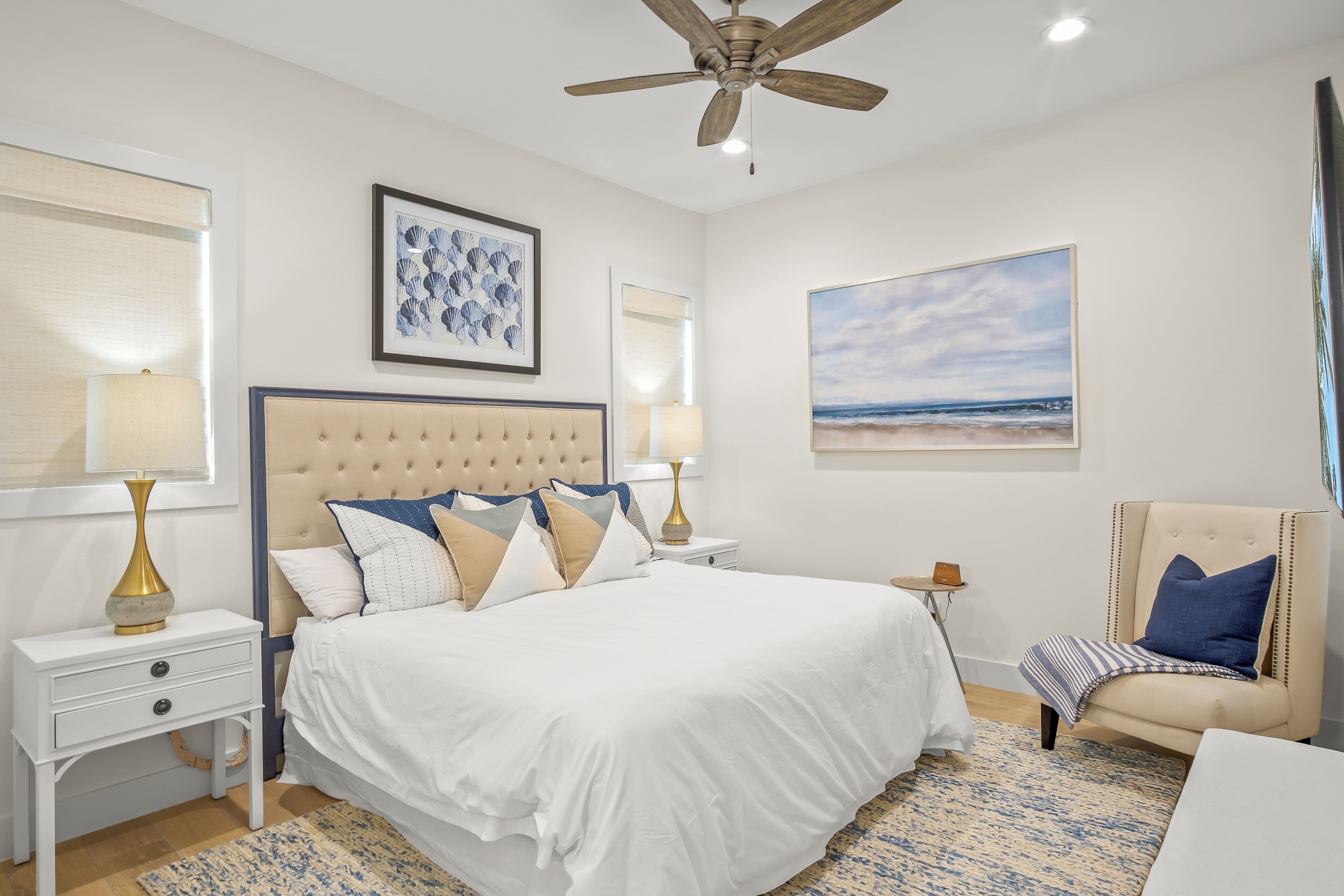 A bedroom with a large bed, a ceiling fan, a nightstand with a lamp, a chair, and a beach-themed painting on the wall.