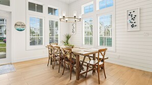 A dining area with a wooden table and chairs, large windows with blinds, and a chandelier. - Beach Blue 227 LAU (Port Aransas)