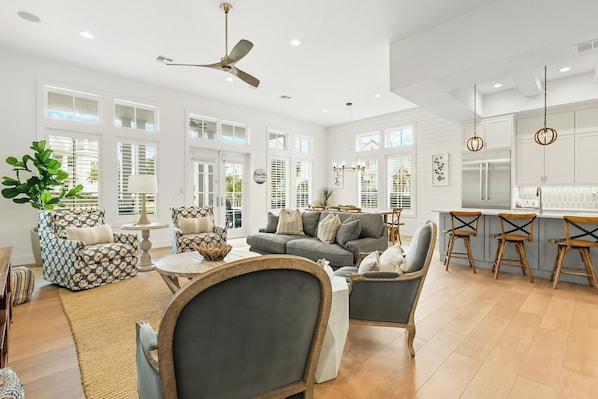 A spacious living room with a gray sofa, patterned armchair, and wooden coffee table. A kitchen area with bar stools and a large window is visible. - Beach Blue 227 LAU (Port Aransas)