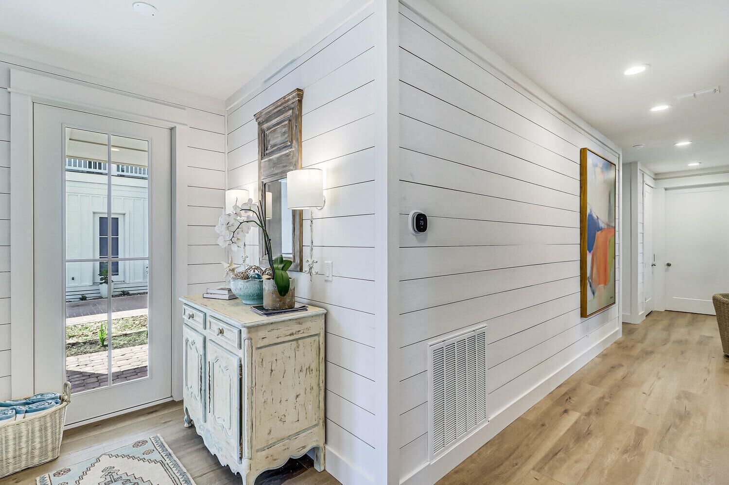 A hallway with white paneled walls, a vintage-style console table with decorative items, a large mirror, and a door leading to an outdoor area.