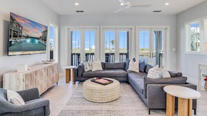 A modern living room with a large TV, comfortable seating, a wooden console, and large windows offering a view of the outdoors. - Surf Camp 104 NEI (Port Aransas)