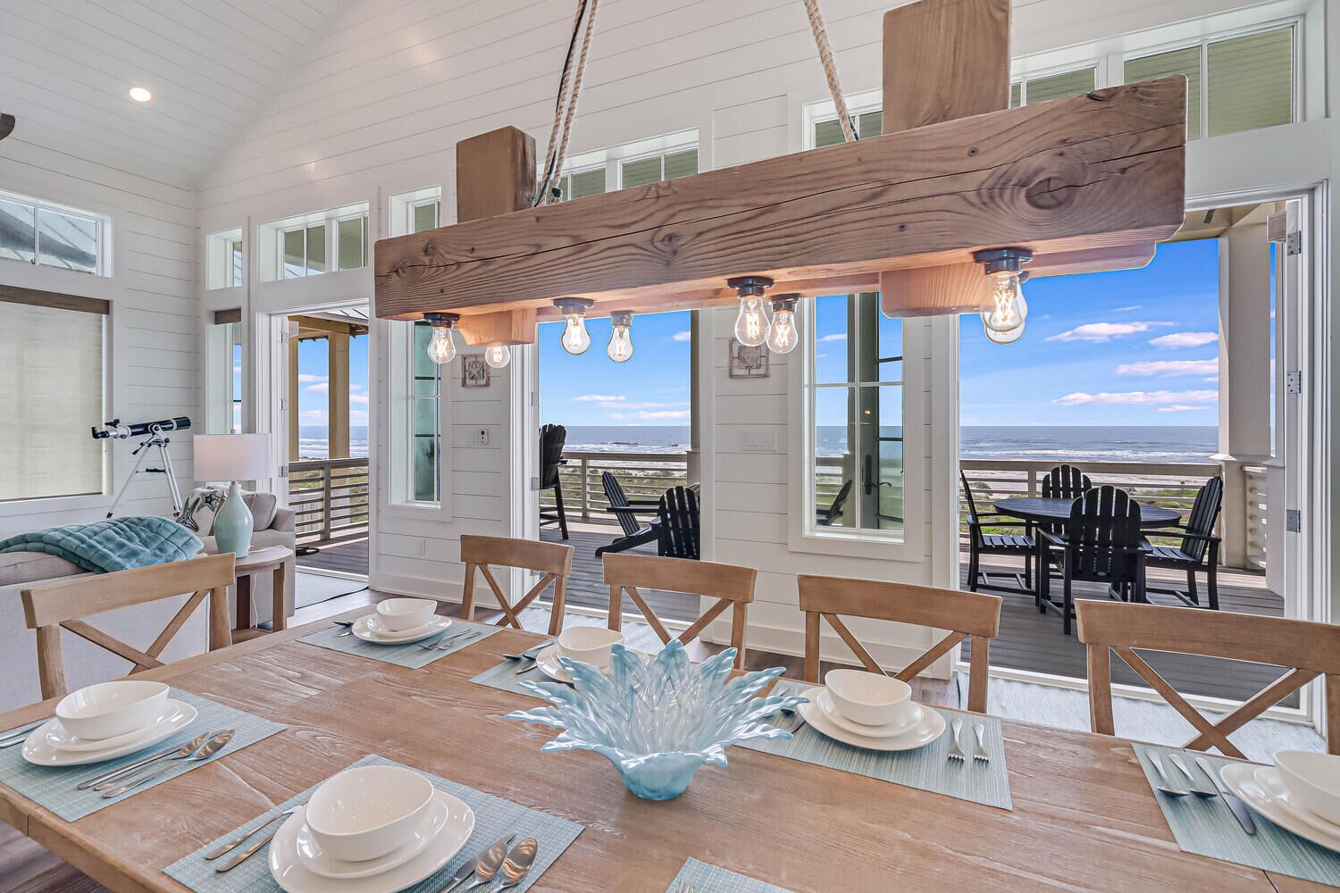 A dining area with a wooden table set for four, large windows, and a view of the ocean.