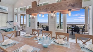 A dining area with a wooden table set for four, large windows, and a view of the ocean. - Livin' the Dream 136 TUC (Port Aransas)