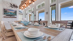 A dining room with a wooden table set for four, large windows, and a view of the ocean. - Livin' the Dream 136 TUC (Port Aransas)