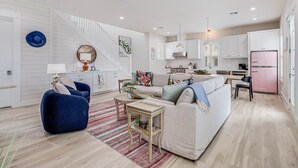 A modern living room with a sofa, coffee table, and blue armchair. A staircase leads to a kitchen with white cabinets and a pink refrigerator. - Rose All Day 220 Esc-c (Port Aransas)