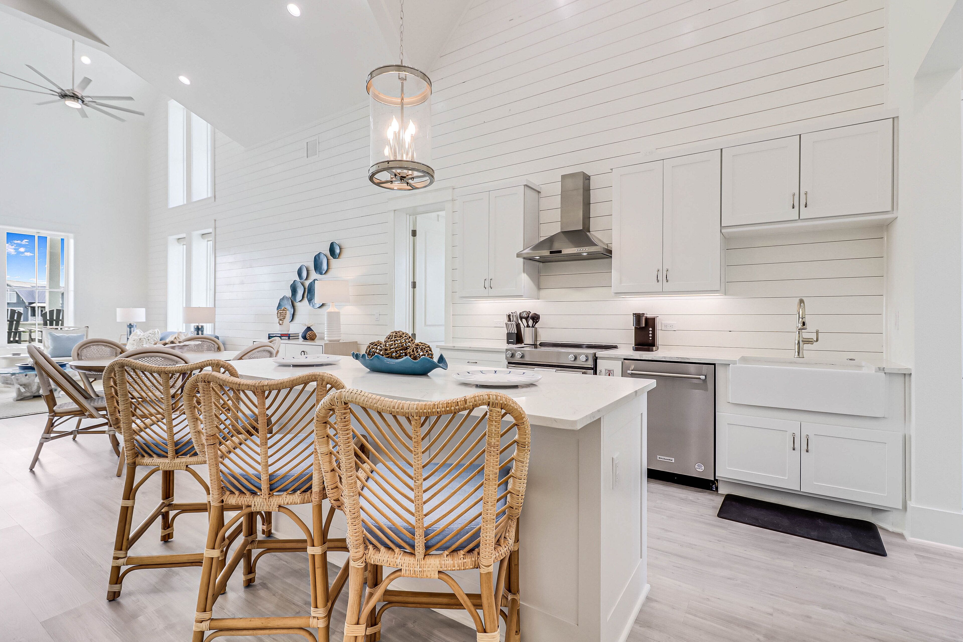 Une cuisine moderne avec des meubles blancs, un îlot central, des chaises en osier et un ventilateur de plafond.