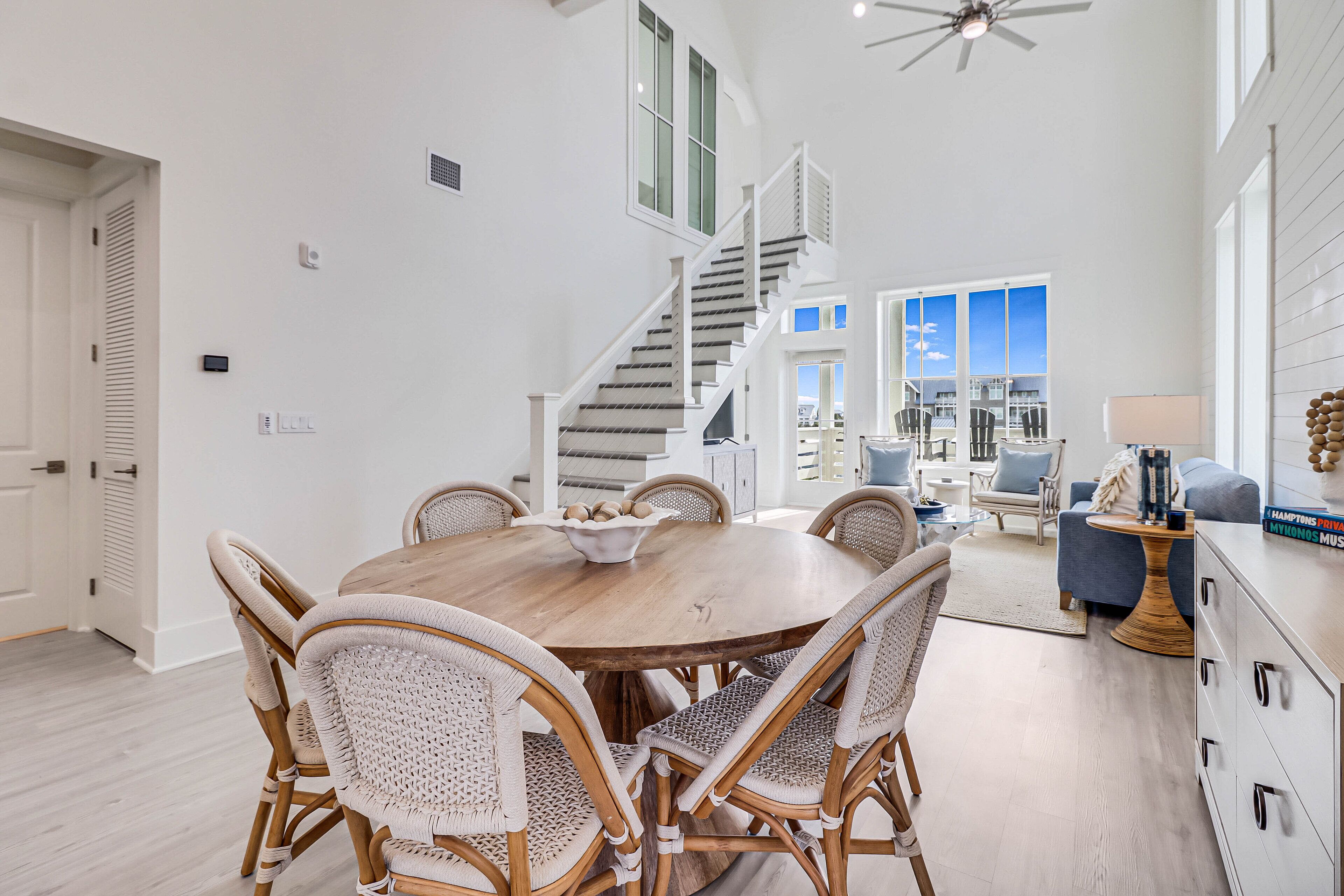A spacious dining area with a wooden table and wicker chairs, a staircase leading to a balcony, and a view of the sky through large windows.