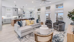 A spacious living room with a gray sofa, a round coffee table, and a gray armchair. There is a ceiling fan, pendant lights, and a large window providing natural light. - Casa Del Sol 213 TEA (Port Aransas)