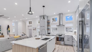 A modern kitchen with a central island, stainless steel appliances, and a grey sofa in the living area. - Casa Del Sol 213 TEA (Port Aransas)