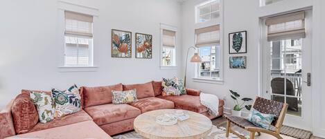 Amplia sala de estar con un sofá modular grande, una mesa de centro redonda y una silla de mimbre. Varias ventanas con persianas, y obras de arte decoran las paredes.