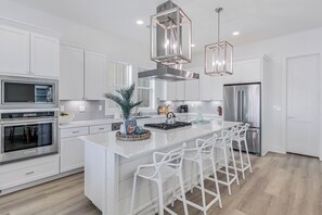 A modern kitchen with white cabinetry, stainless steel appliances, a central island with bar stools, and pendant lighting. - Latin Vibe 312 SOU (Port Aransas)