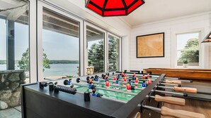 A foosball table in a room with large windows overlooking a body of water, a stone wall, and a wooden bench.