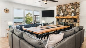 A spacious living area with a large wooden table, grey upholstered sofas, a ceiling fan, a stone wall, and a TV mounted on the wall.