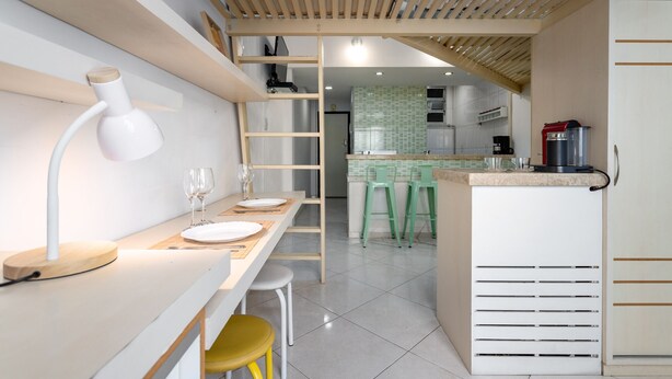A modern kitchen with a white countertop, a built-in stove, and a coffee machine. There is a dining area with a white table and chairs, and a wooden ladder leading to a lofted space.