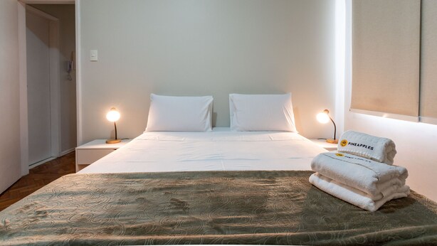 A neatly made bed with white linens and two pillows, a bedside lamp, and a folded towel with a pineapple logo on a bedside table.