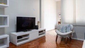 A modern living room with a flat-screen TV on a white entertainment unit, a wooden herringbone floor, and a light-colored armchair.