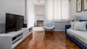 A modern living room with a flat-screen TV on a white entertainment unit, a blue sofa with a white throw blanket, and a herringbone wood floor.