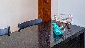 Una cocina con encimera negra, una lámpara colgante de cobre, una cesta tejida con follaje y una escultura de pájaro color turquesa.