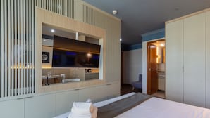 Una habitación de hotel moderna con una cama grande, un televisor de pantalla plana montado en un panel de madera, una pequeña kitchenette y un baño visible a través de una puerta abierta.