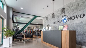 Un mostrador de recepción con acabado de madera y un panel negro, una pared de mármol con el texto "MUND NOVO" y un logotipo de globo, luces colgantes y una escalera al fondo.