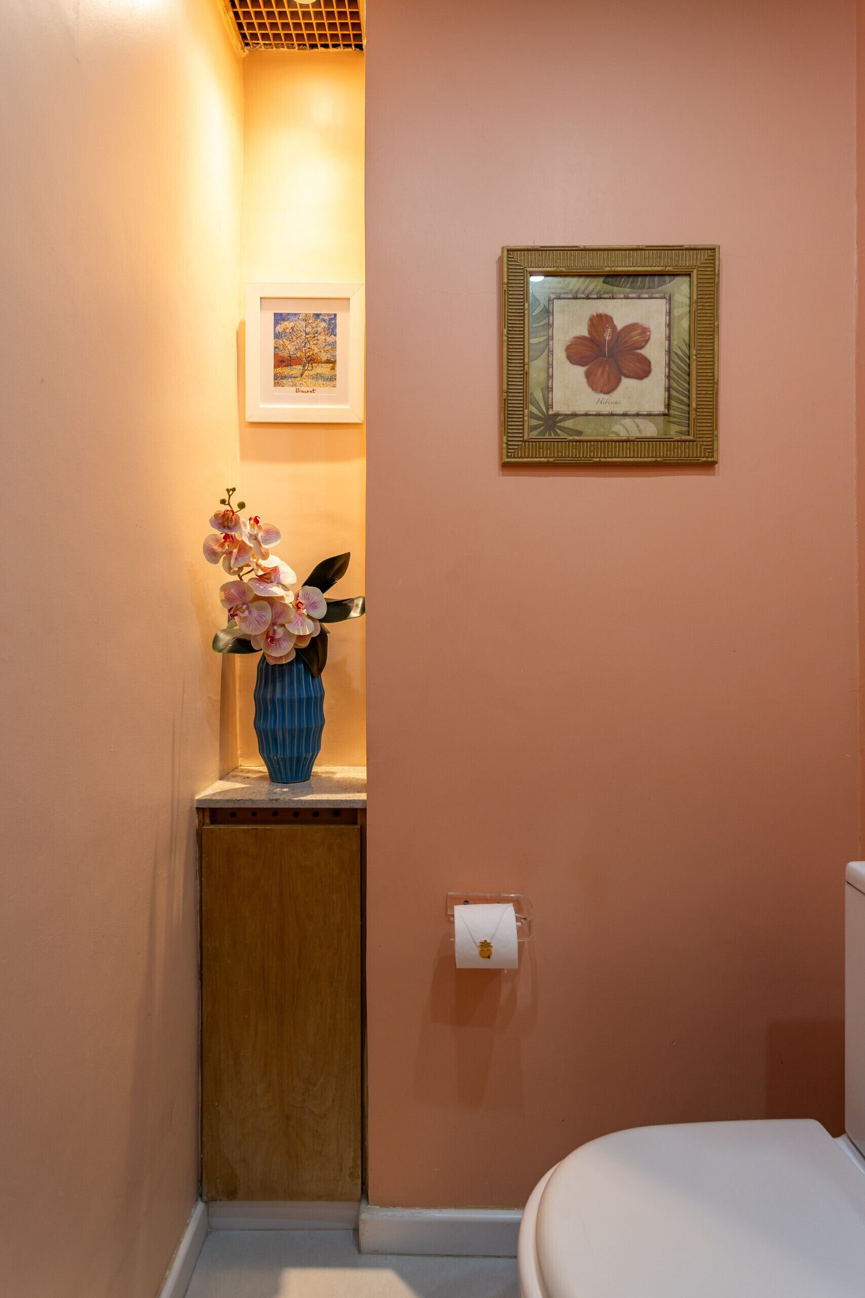 Een badkamer met een toilet, een fotokader met een hibiscusbloem en een vaas met orchideeën op een klein plankje.