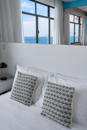 A hotel room with a bed, two patterned pillows, a black lamp, and a view of the sea through large windows.