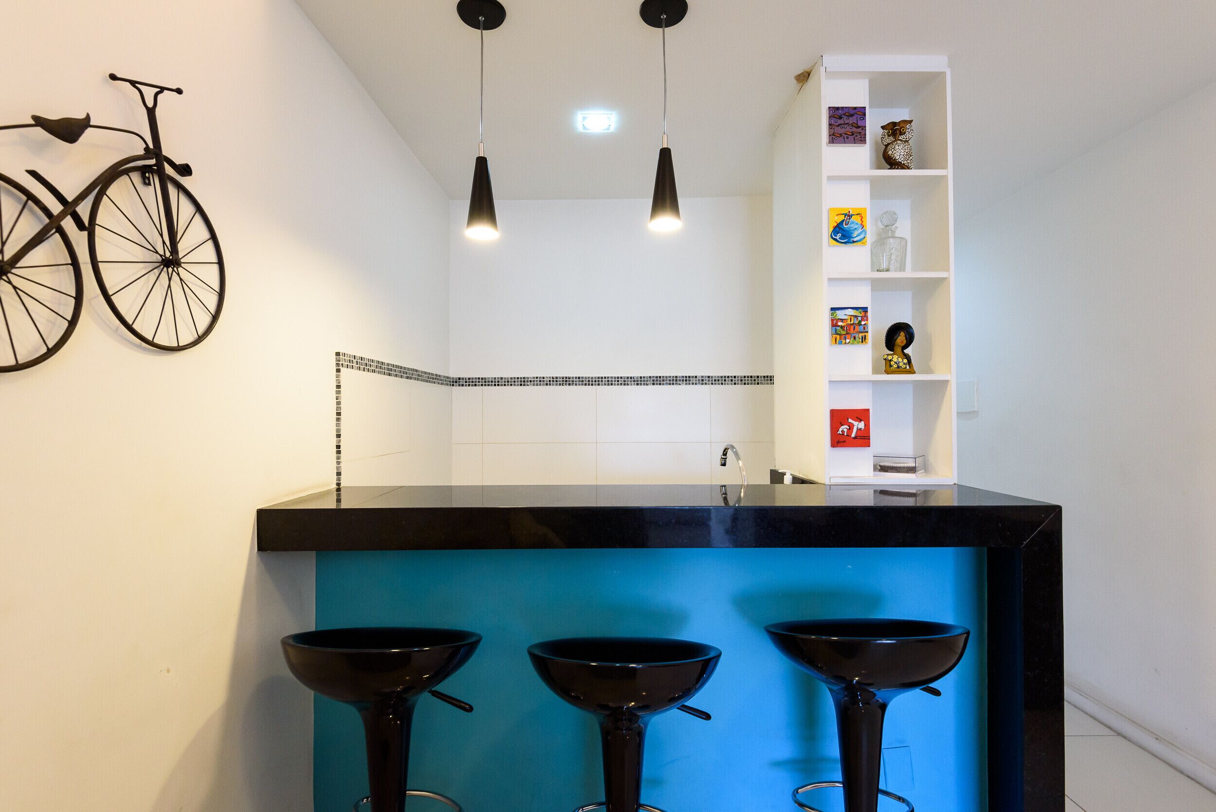 Una cocina con encimera negra, tres taburetes negros, una bicicleta en la pared y un estante blanco con objetos decorativos.