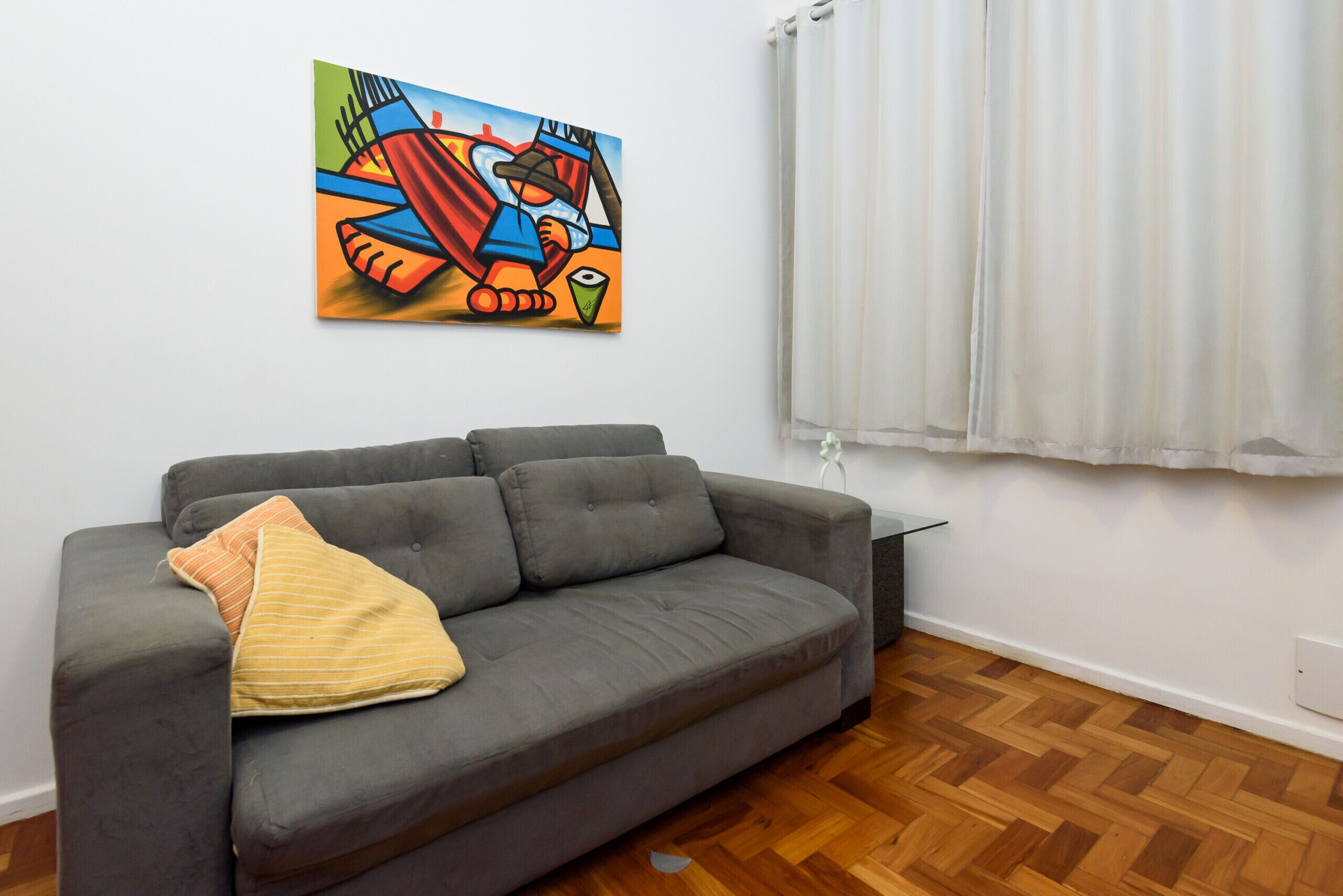 Un sofá gris con un cojín a rayas, un cuadro abstracto de colores en la pared y una ventana con cortinas blancas.