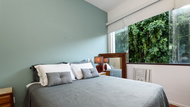 A bedroom with a bed, a nightstand, a mirror, and a window with a view of greenery. - Peace in Copacabana | Forest view | BR1009 (Rio de Janeiro)