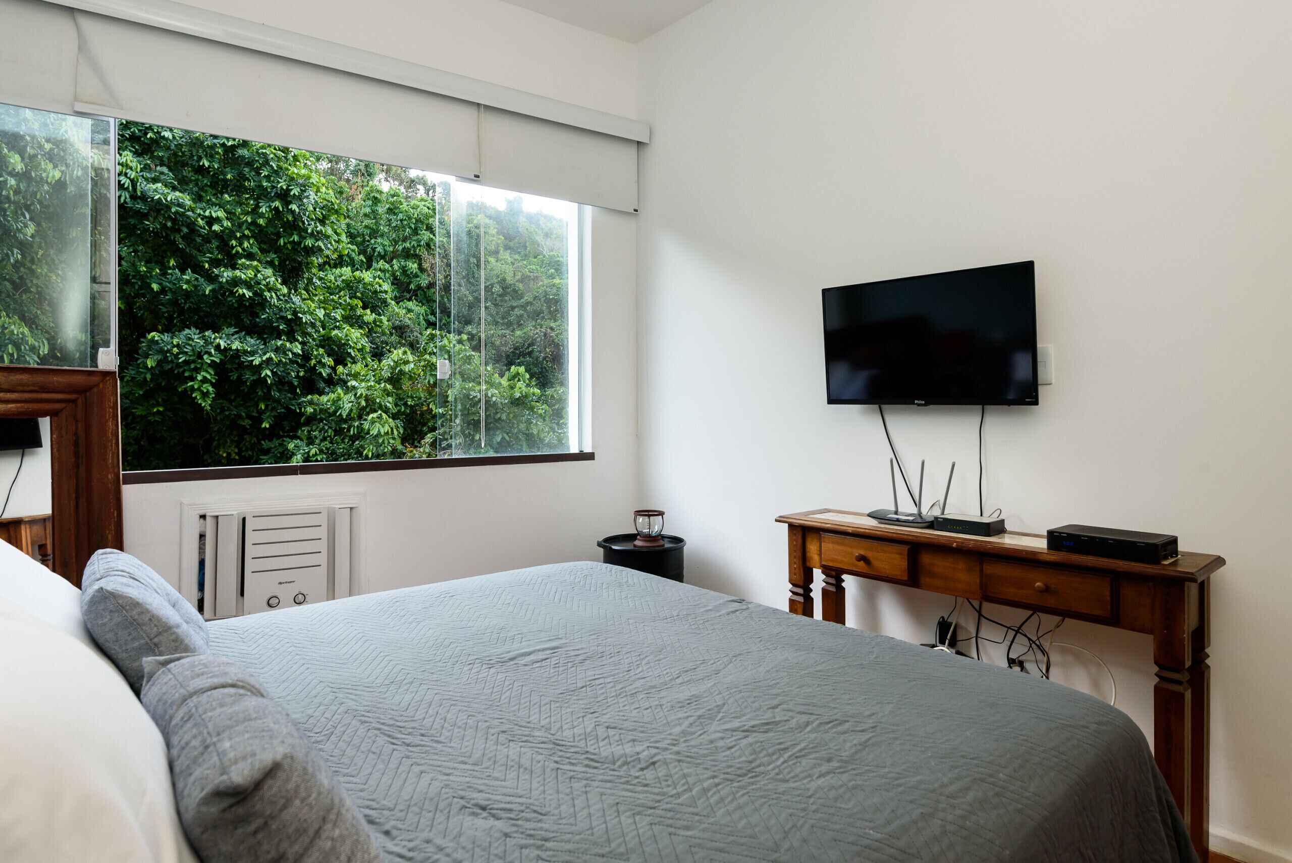 A bedroom with a large bed, a TV mounted on the wall, a wooden desk, and a window with a view of greenery.