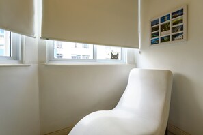 A white chair in a corner of a room with a window and a wall-mounted picture. - Side sea view, 3min from the beach | PJ204 (Rio de Janeiro)