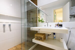 A modern bathroom with a glass shower enclosure, a white sink with a mirror above, and a small shelf with toiletries. - Side sea view, 3min from the beach | PJ204 (Rio de Janeiro)