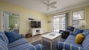 Een woonkamer met gestreepte blauwe banken, een witte salontafel, een plafondventilator, een televisie op een witte kast, en een raam met jaloezieën.