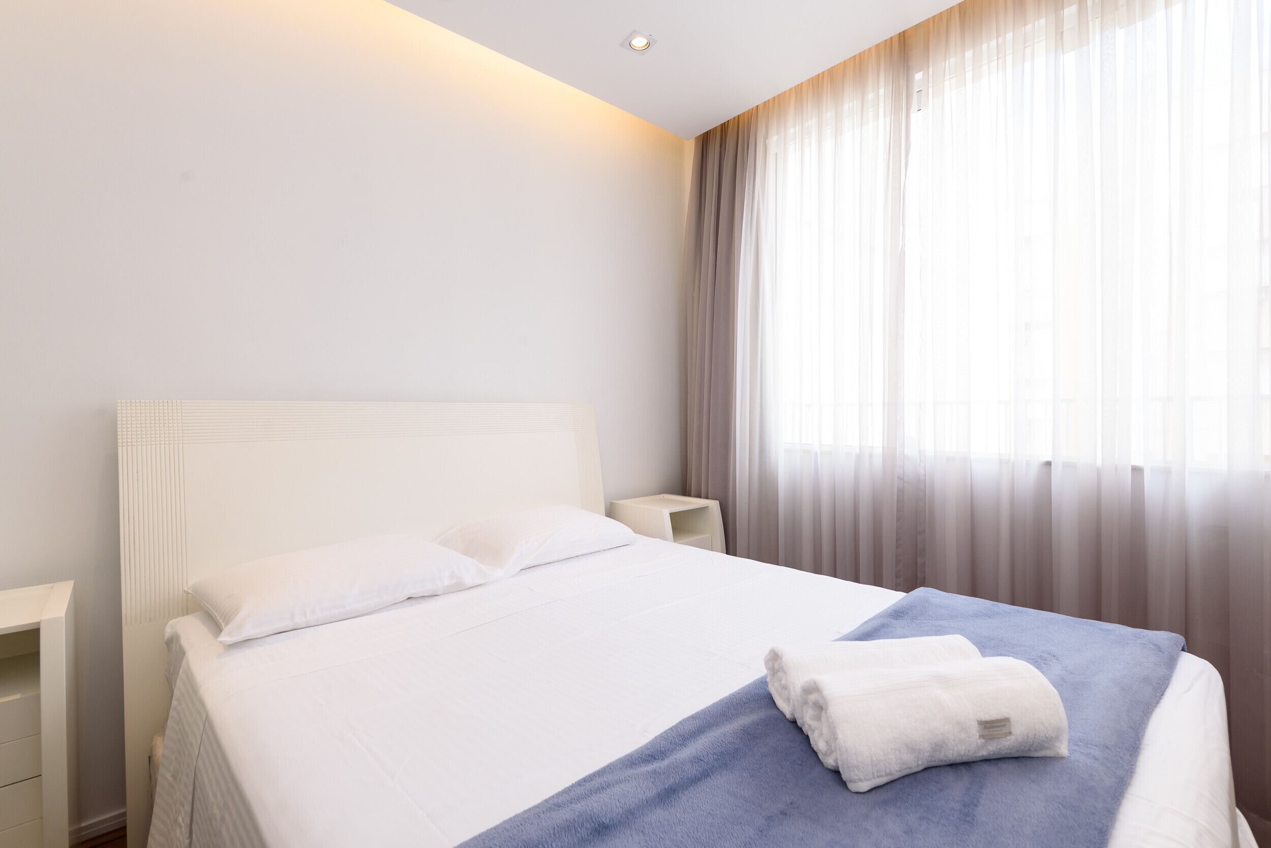 Una cama bien hecha con sábanas blancas y una colcha azul, una mesita de noche y una ventana con cortinas traslúcidas.