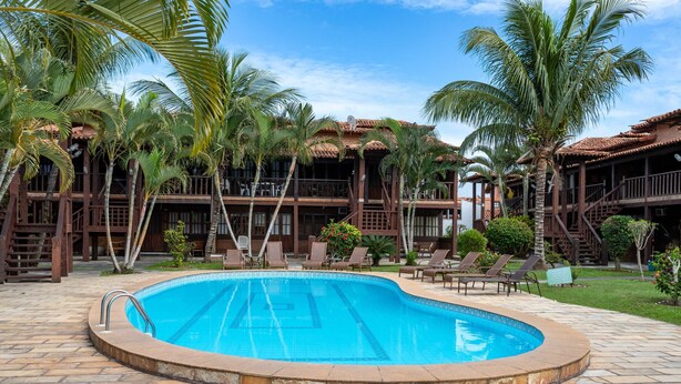 A swimming pool surrounded by palm trees and a wooden building with balconies. - Apart Hotel  | BUZ215 (Armação dos Búzios)