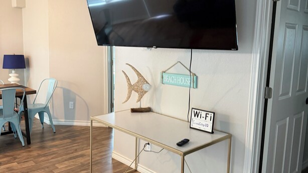 A modern living room with a flat-screen TV mounted on the wall, a small table with a sign indicating Wi-Fi availability, a colorful rug, and a door leading to another room.